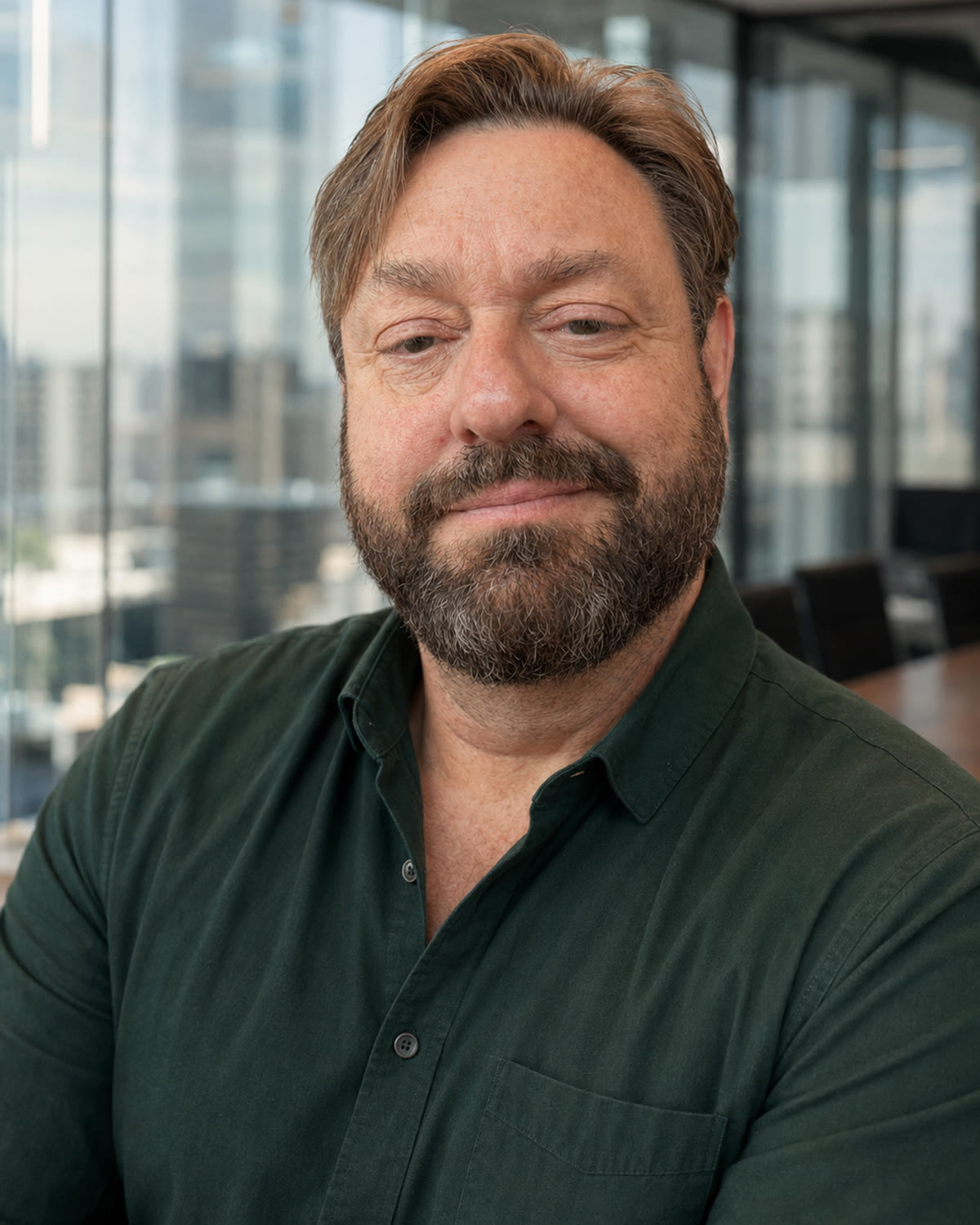 Guy Flexor, a middle-aged man with a salt-and-pepper beard, in a forest-green shirt, looking at the camera in a bright office with a city skyline behind him.